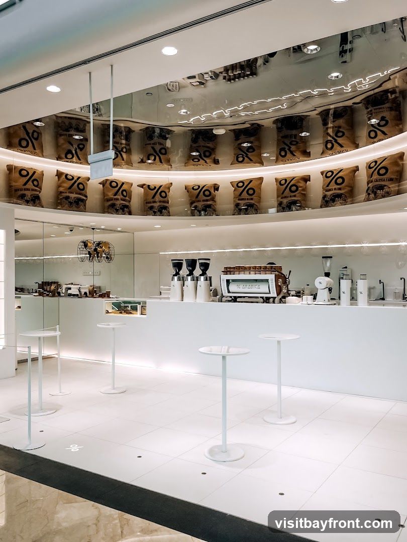A clean and minimalist coffee shop interior with white walls and floors. Several tall, narrow tables with round tops are placed around the seating area. Behind the counter, a professional espresso machine and coffee grinders are visible. The shelves above the counter are lined with burlap sacks featuring a prominent percentage logo, suggesting coffee bean packaging. The ceiling is a reflective surface, mirroring the display of sacks and lights.