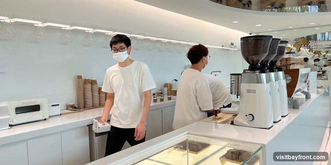A bright and modern coffee shop interior with baristas preparing drinks. The counter features coffee grinders, cups, and a toaster. Decorative globes and bags are visible in the background.