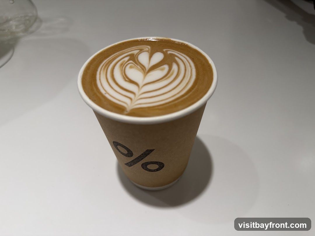 A close-up shot of a latte in a paper cup with the "%" logo. The latte has intricate latte art on top.