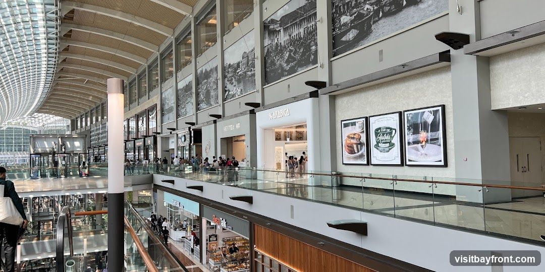 The image shows the interior of a multi-level shopping mall with a distinctive curved ceiling and large windows. Several shops are visible on different levels, including % ARABICA and HEYTEX. Large black and white murals depicting historical scenes adorn the upper walls. A glass railing separates the walkway from the lower levels. People are seen browsing and walking around the mall.