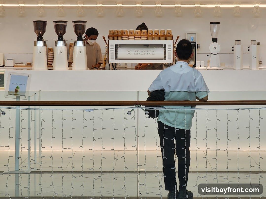 An interior view of a modern coffee shop with baristas preparing drinks behind a counter. The counter features coffee grinders, an espresso machine, and cups. A sign indicates "% ARABICA" on the espresso machine. A small sign advertises "MATCHA SOFT CREAM". A person stands at the counter, facing away from the camera. The background has decorative lights.