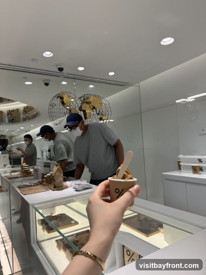 A person holds a cup of soft serve ice cream with a wooden spoon. The cup has a logo with two percent signs. In the background, baristas are working behind a counter in a modern, minimalist coffee shop. The counter has a display case with pastries or desserts. Two large globe decorations hang from the ceiling. The walls are white, and there are security cameras visible on the ceiling.