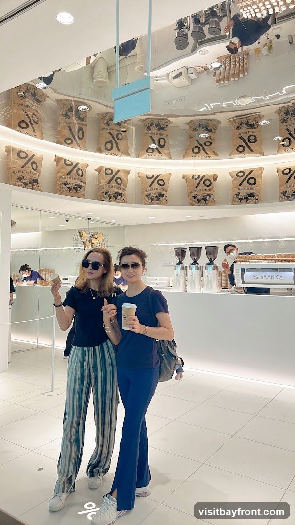 Two women are standing in a brightly lit, modern cafe, holding coffee cups. The cafe features a minimalist design with white walls and a reflective ceiling. Behind the counter, bags with the "% ARABICA" logo are displayed on a curved shelf. The counter itself has coffee machines and a sign that reads "% ARABICA".