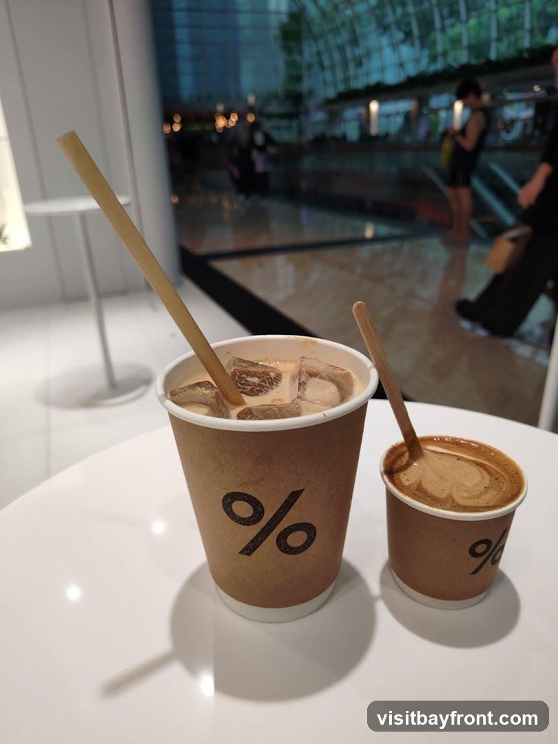 Two disposable coffee cups with the '%' logo are on a white table. The larger cup contains an iced coffee with a straw, and the smaller cup contains a hot coffee with latte art and a wooden stirrer. In the background, a blurred view of a modern interior with people and escalators is visible.