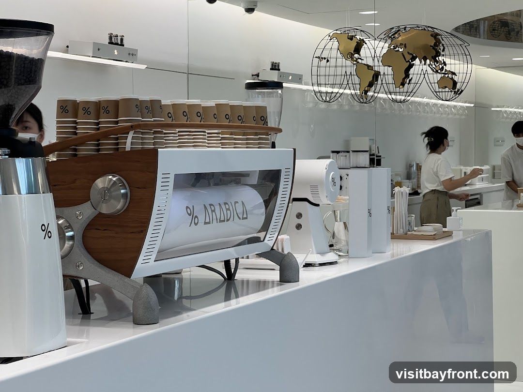 A modern coffee shop interior with a prominent espresso machine, stacks of branded paper cups, and a world map decoration on the wall. Baristas are visible in the background preparing drinks. The overall aesthetic is clean and minimalist with white and wood accents.