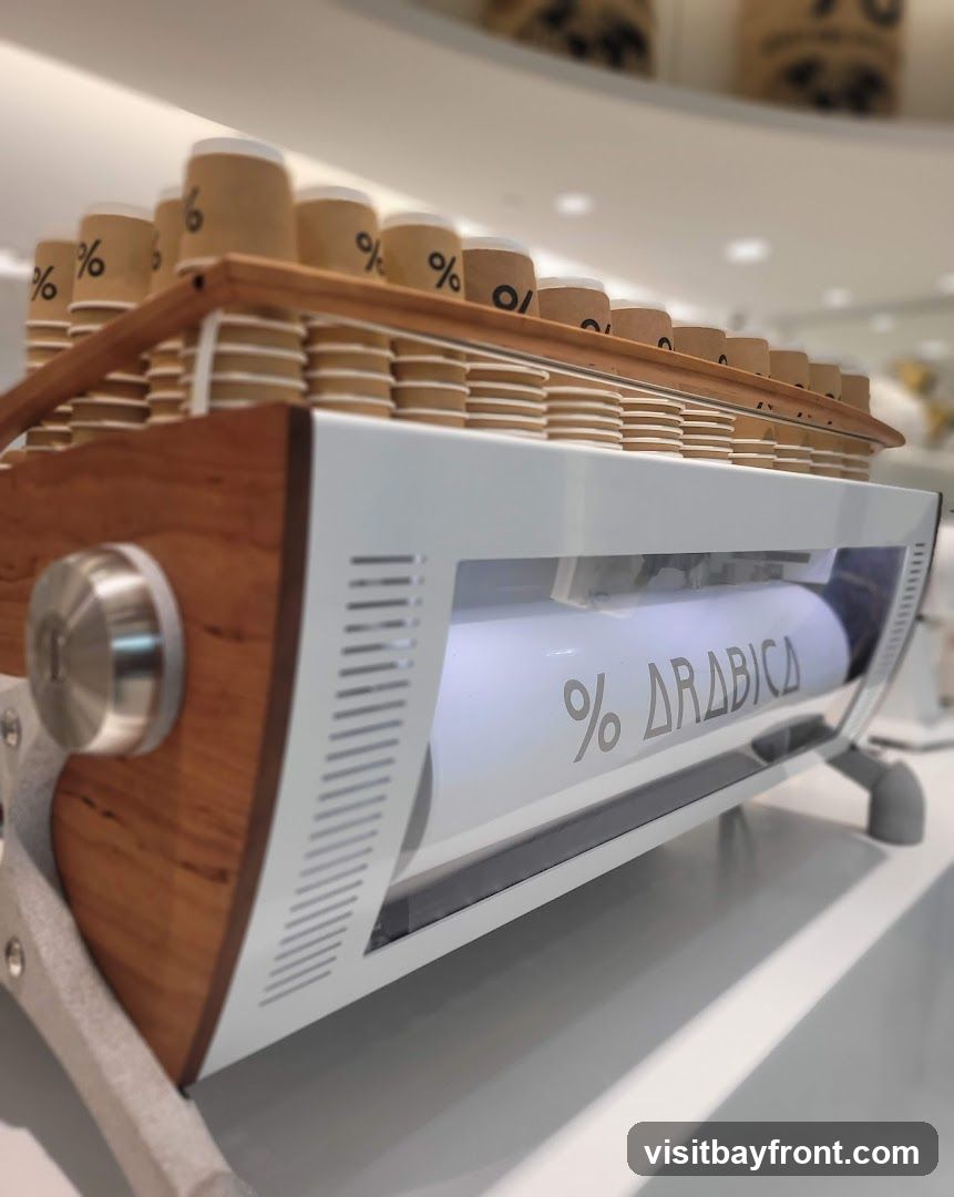 A close-up, low-angle shot of a modern coffee machine. The machine features a white exterior with wooden accents and a clear glass panel. Inside the glass panel, a white cylinder is visible with the text "% ARABICA" etched onto it. Stacked on top of the machine are numerous disposable coffee cups, each with the "%" logo printed on them. The background is blurred, suggesting an indoor setting, possibly a cafe.