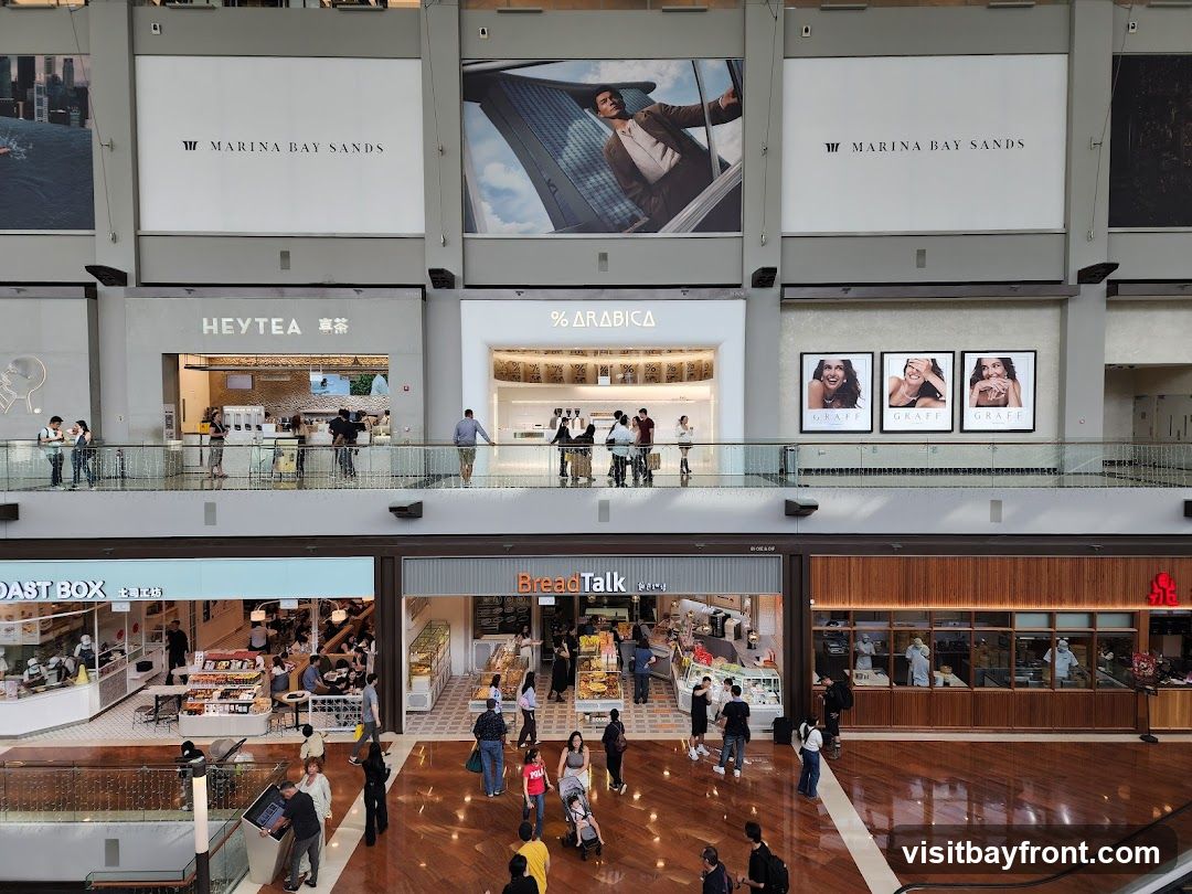 An interior view of a shopping mall with multiple storefronts visible on two levels. On the upper level, signs for Marina Bay Sands, Heytea, and % Arabica are prominent. On the lower level, storefronts for Roast Box and BreadTalk are visible, along with other shops. People are seen browsing and walking around the mall.