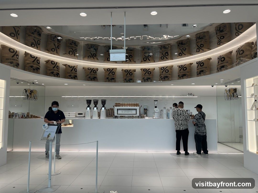 A modern coffee shop interior with a white counter and shelves displaying coffee bags. Several customers and staff are visible. The ceiling features a curved display of coffee bean bags with the percentage logo.