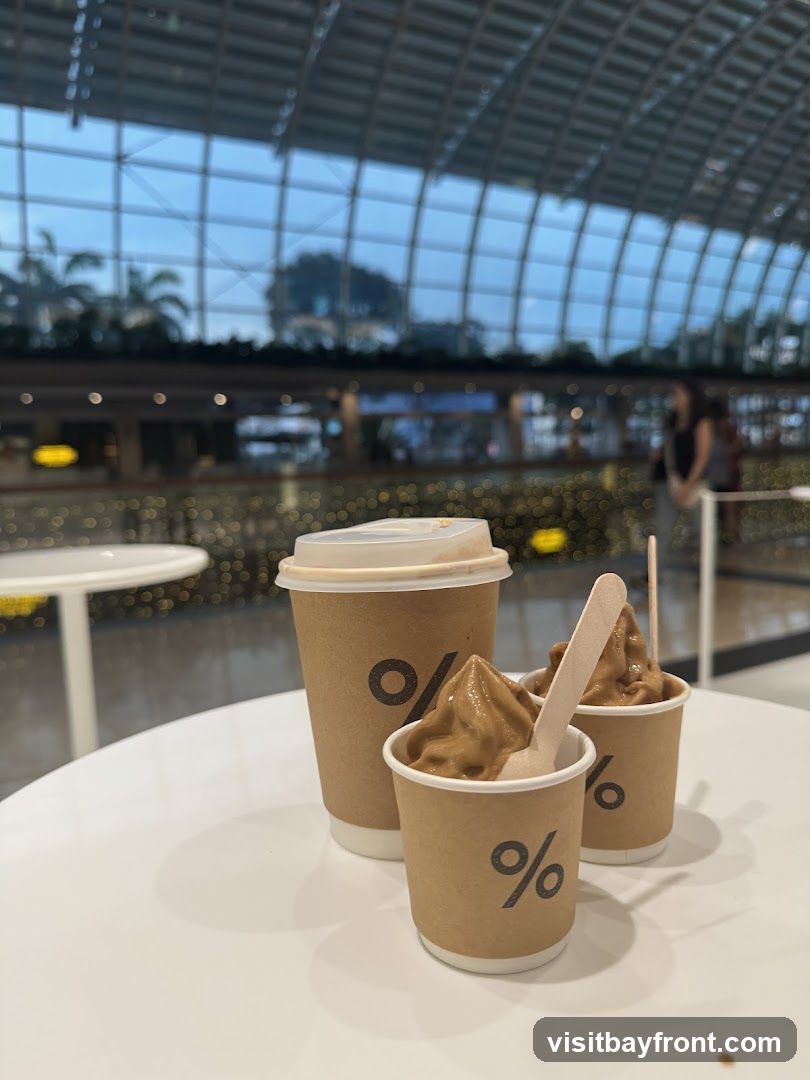 A large coffee cup and two small cups of soft serve ice cream are on a white table. The cups have a percentage logo. In the background, a blurred view of a modern building interior with a glass ceiling and lights is visible.