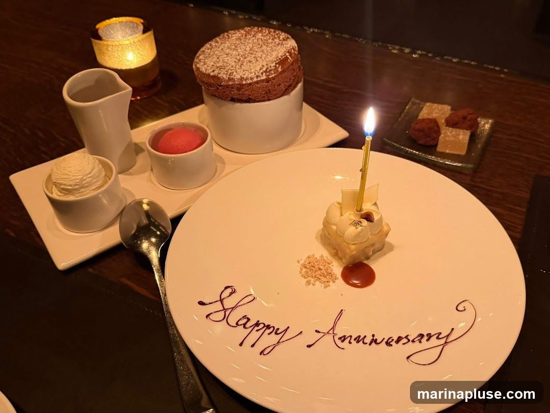 A white dessert plate with "Happy Anniversary" written in chocolate sauce. In the center of the plate is a small, square dessert topped with cream, a white chocolate shard, and a gold leaf, with a lit candle. To the left of the plate are three small white bowls containing ice cream, a scoop of sorbet, and a pitcher of sauce. Behind these is a chocolate soufflé. In the background, a small glass dish holds two pieces of candy. A lit candle in a decorative holder is also visible.
