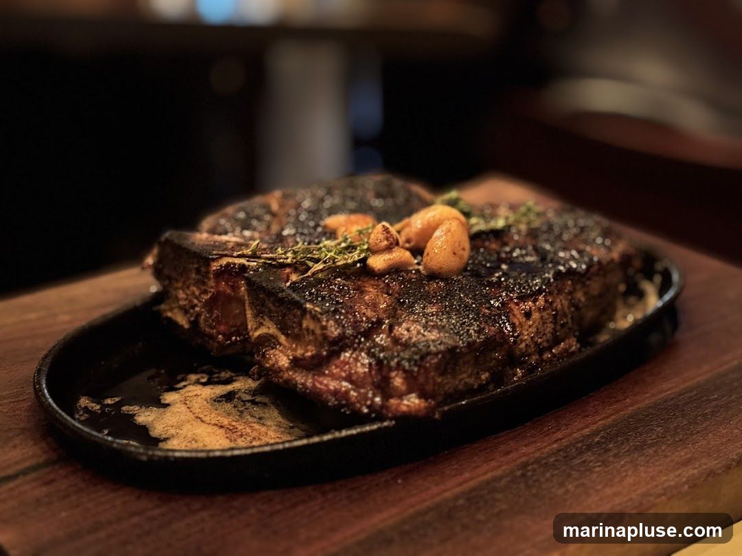 A thick, seared T-bone steak is presented on a cast-iron platter, garnished with roasted garlic cloves and fresh herbs. The steak has a dark, crusty exterior and appears to be served with melted butter or pan juices. The platter rests on a wooden surface, with a blurred background suggesting a restaurant setting.