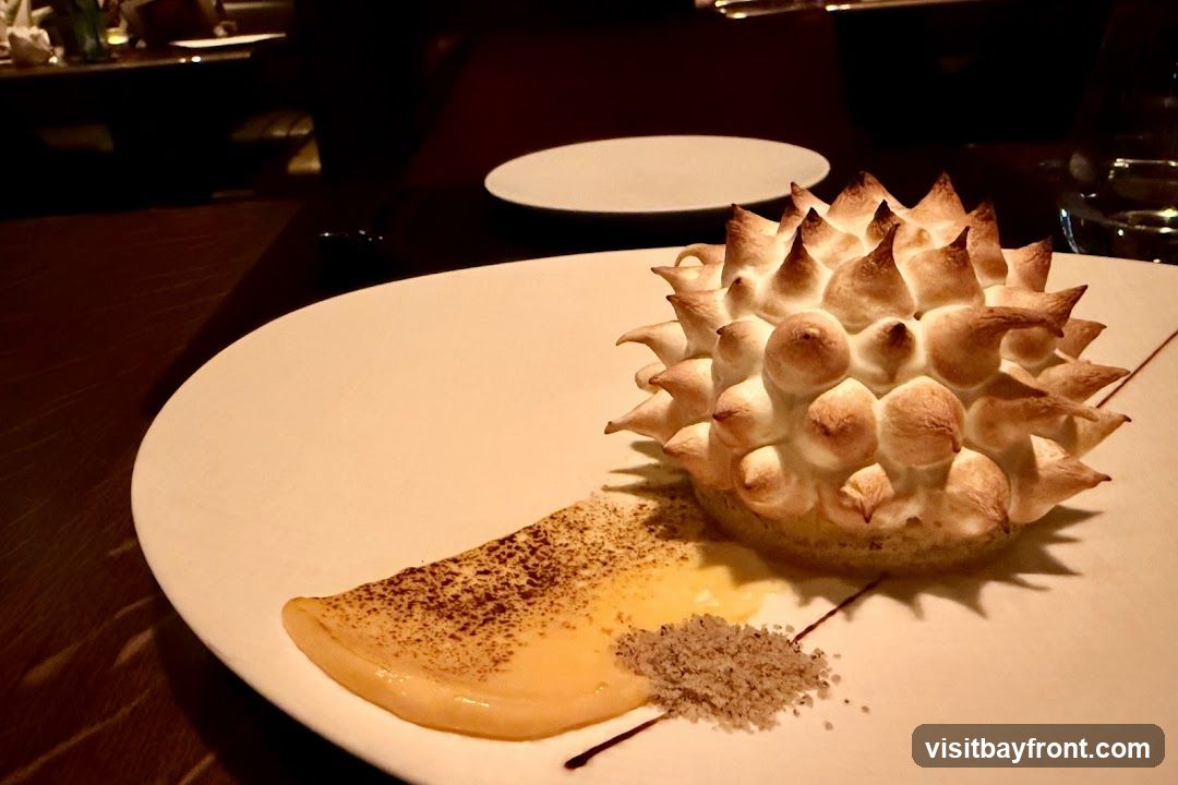 A close-up shot of a dessert on a white plate. The dessert is a tart with a spiky, toasted meringue topping. It is served with a dollop of yellow custard and a sprinkle of coarse sugar. The background is dark and out of focus, suggesting a restaurant setting.