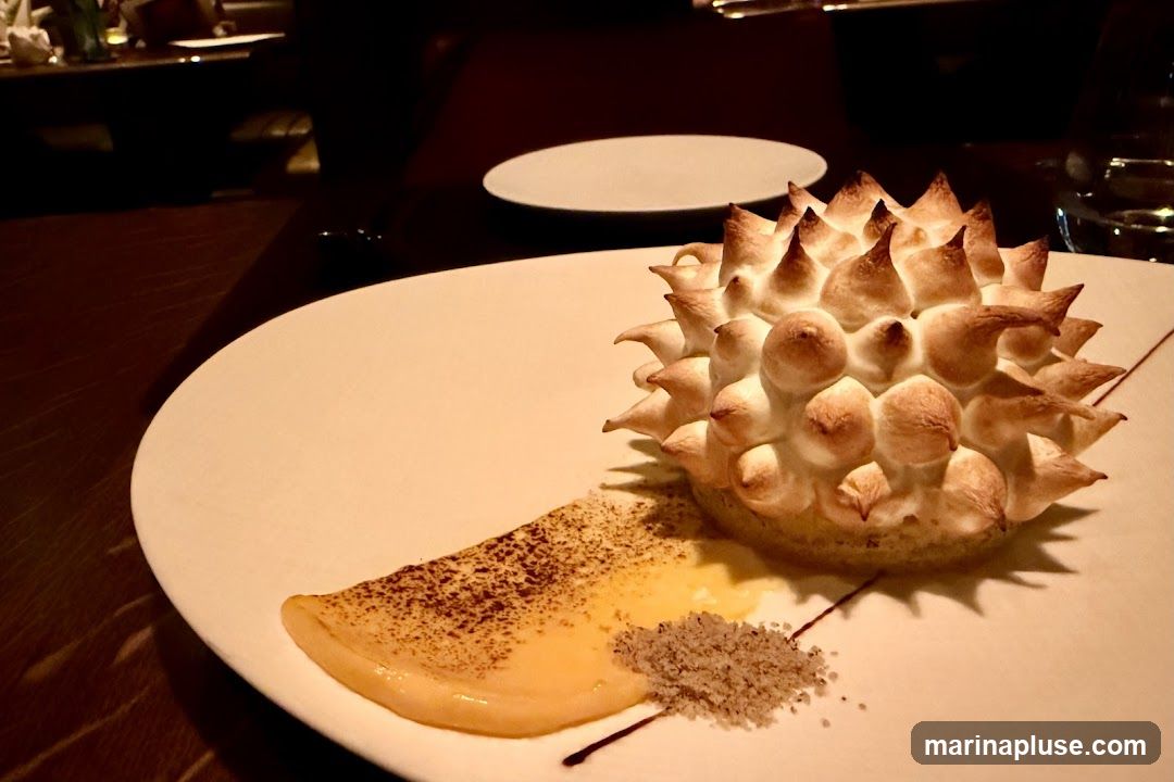 A close-up shot of a dessert on a white plate. The dessert is a tart with a spiky, toasted meringue topping. It is served with a dollop of yellow custard and a sprinkle of coarse sugar. The background is dark and out of focus, suggesting a restaurant setting.