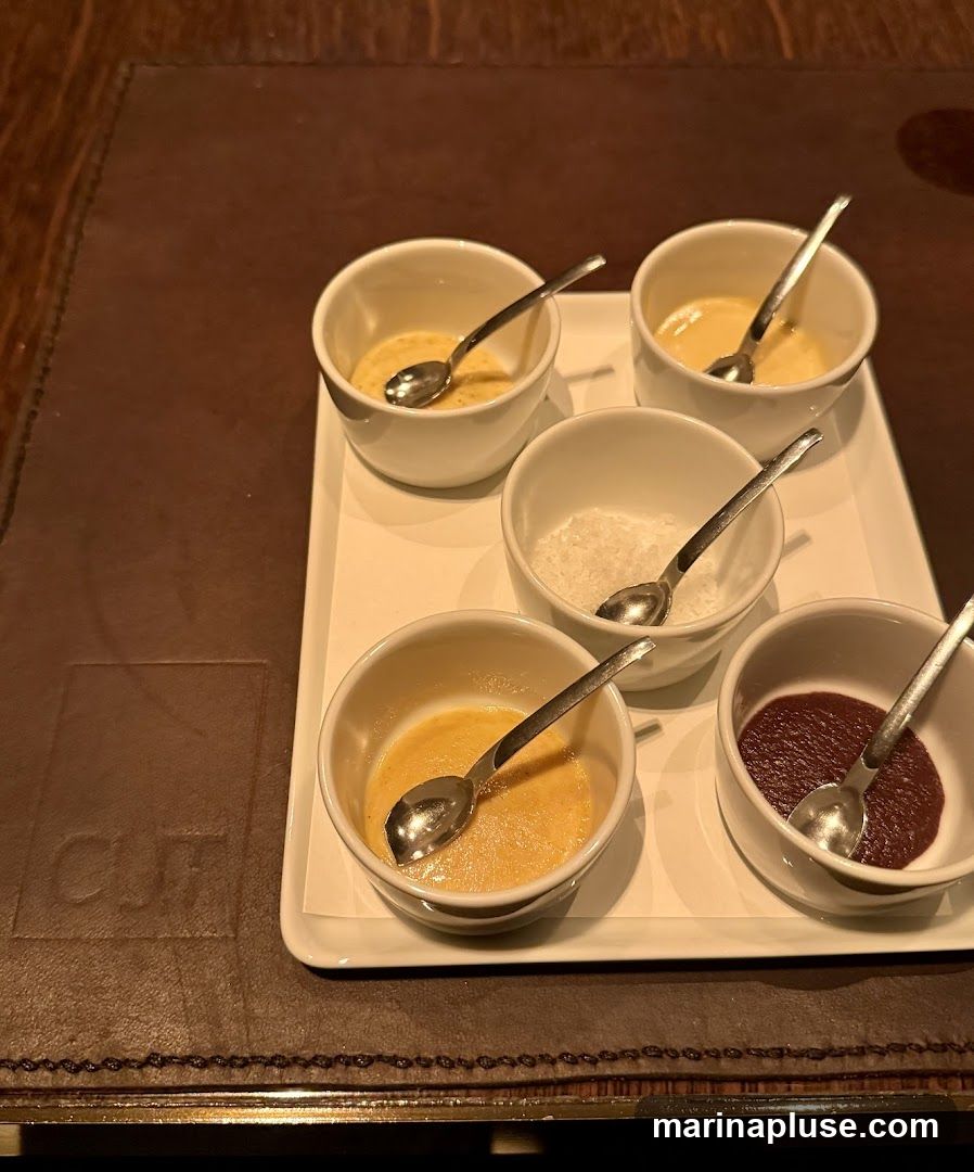 A white tray holds five small white bowls, each with a spoon. Three bowls contain a light yellow, creamy substance. One bowl contains white granular material, possibly salt. The fifth bowl contains a dark red, thick sauce. The tray is placed on a dark brown textured surface, possibly a placemat, with a faint embossed logo that appears to read "CLIF".