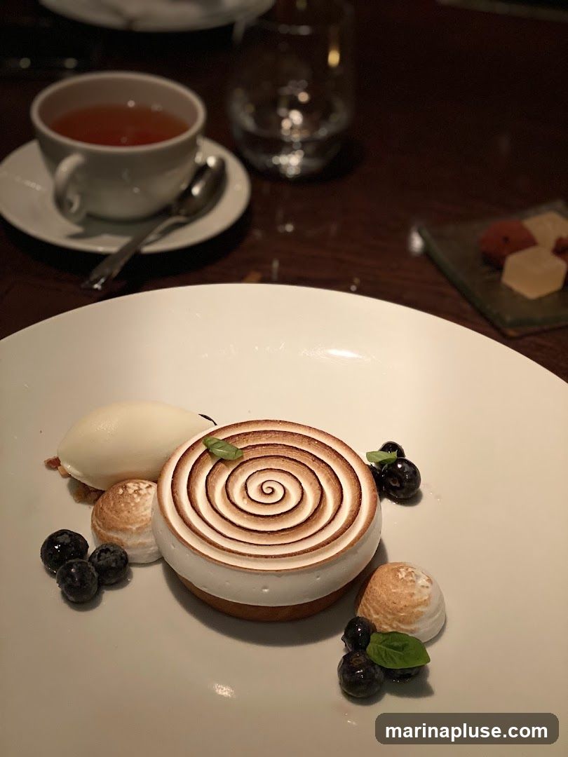 A close-up shot of a beautifully presented dessert on a white plate. The dessert features a circular tart topped with torched meringue in a spiral pattern, accompanied by a scoop of ice cream, several blueberries, and small meringue kisses. A cup of tea and a glass of water are visible in the blurred background, along with a small dish of what appears to be candies or fruit pieces.