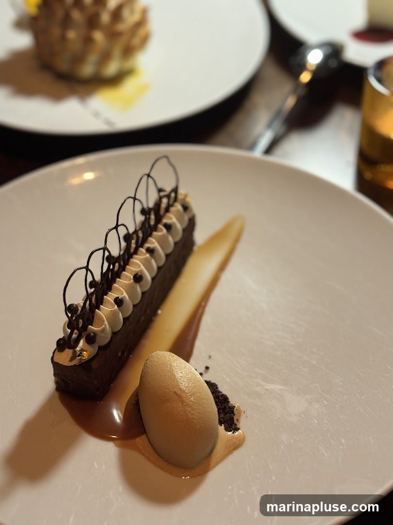 A close-up shot of a chocolate dessert on a white plate. The dessert is a rectangular chocolate cake topped with white frosting and intricate chocolate decorations. It is served with a scoop of ice cream and a drizzle of caramel sauce. Another dessert is visible in the blurred background.