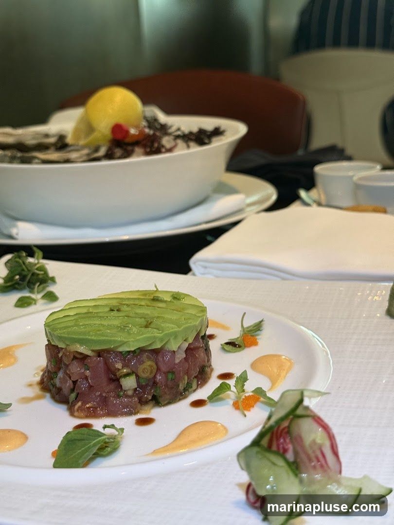 A close-up shot of a finely diced tuna tartare dish topped with sliced avocado, served on a white plate with dollops of sauce and garnished with microgreens. In the background, a bowl of oysters with a lemon wedge and garnish is visible, along with white napkins and cups.