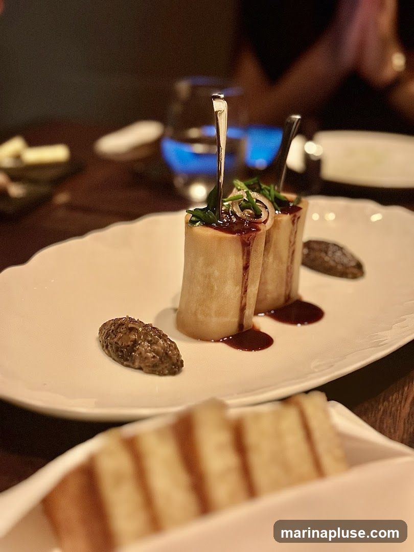 A close-up shot of two pieces of roasted bone marrow served on a white oval plate. The bone marrow is topped with a dark sauce and garnished with fresh herbs and thinly sliced onions. Two small dollops of a dark, textured paste are placed on either side of the bone marrow. In the background, a glass with a blue flame is visible, along with other blurred dishes and diners. In the foreground, a white dish filled with sliced bread is out of focus.
