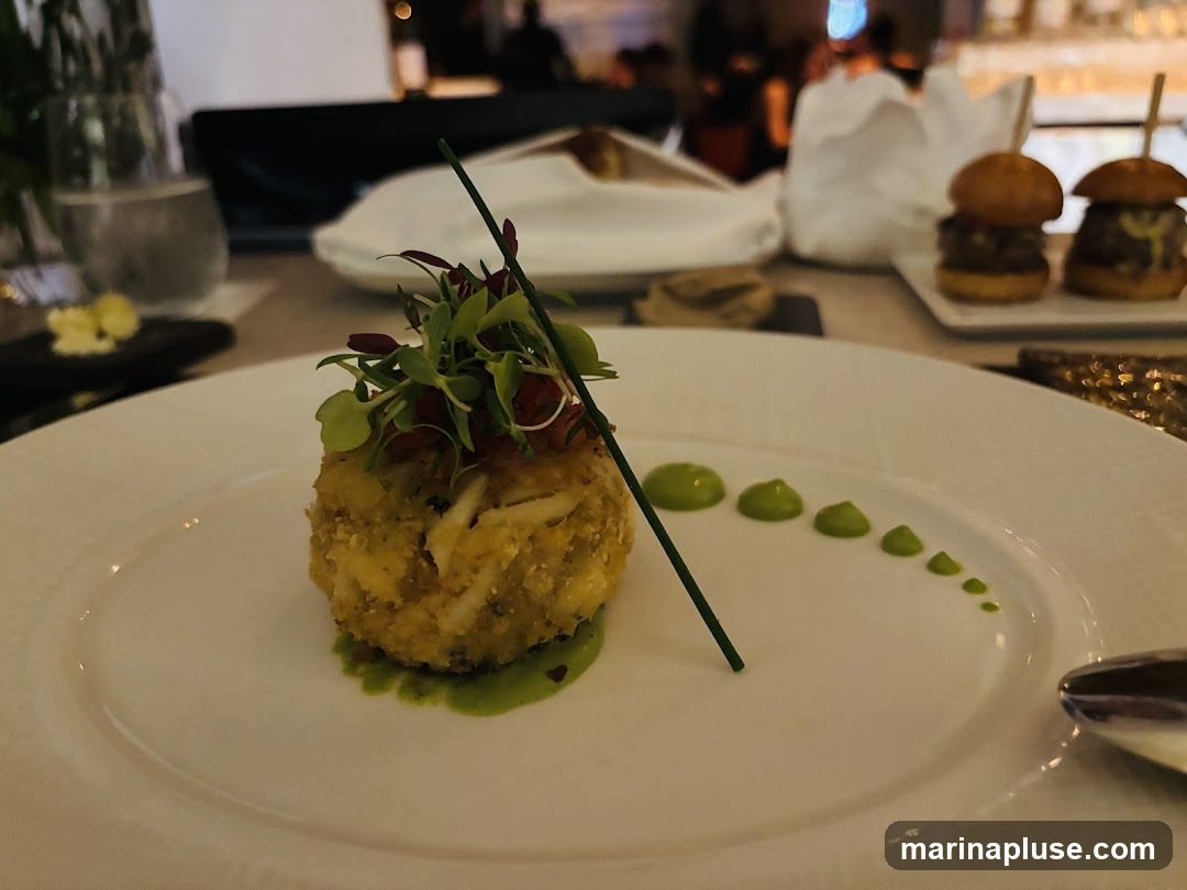 A close-up shot of a crab cake appetizer on a white plate. The crab cake is topped with microgreens and a thin skewer. Green sauce is artfully dotted on the plate. In the background, two mini burgers are visible on a platter, along with a glass of water and other table settings. The lighting suggests an indoor dining environment.