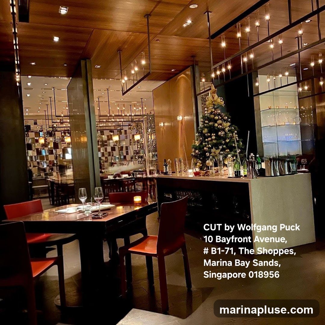 The image shows the interior of a restaurant with a dining table set for two in the foreground. A Christmas tree decorated with ornaments and a gold bow is visible on the right side of the image, near a bar area. The restaurant has a modern and elegant design with wooden ceilings and ambient lighting. The text overlay provides the name and address of the establishment: CUT by Wolfgang Puck, 10 Bayfront Avenue, # B1-71, The Shoppes, Marina Bay Sands, Singapore 018956.