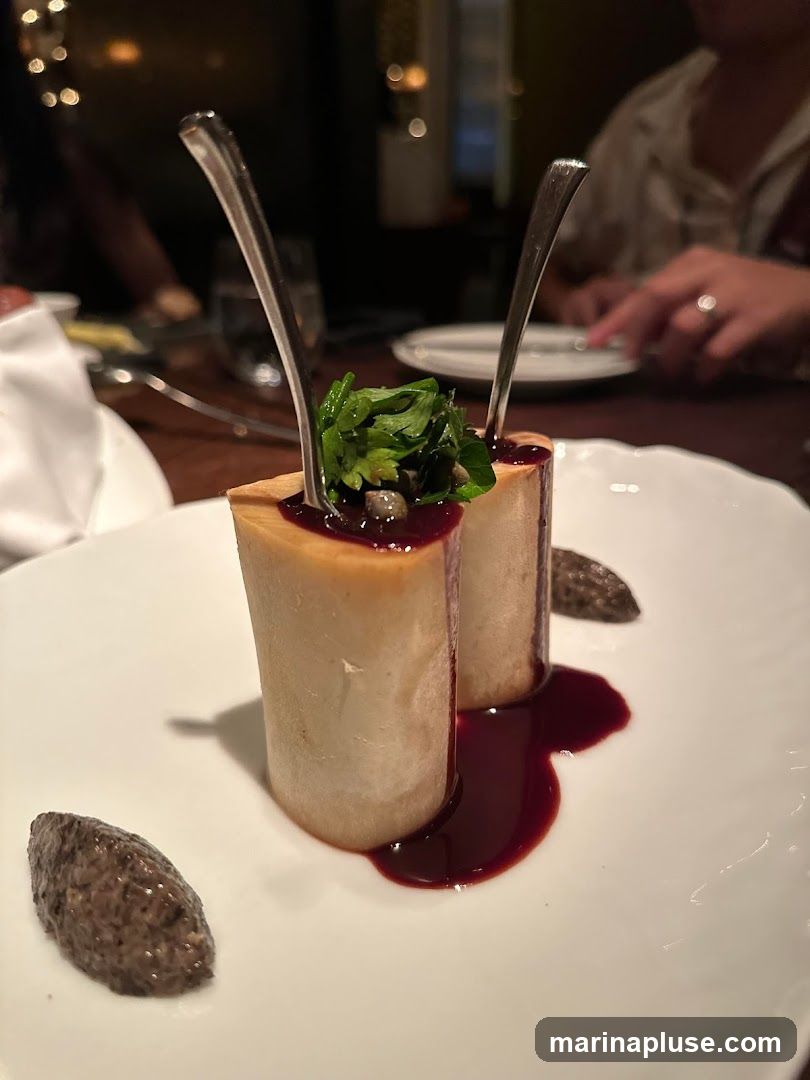 A close-up shot of two pieces of roasted bone marrow, each topped with fresh herbs and capers, served on a white plate with a dark red sauce and a side of what appears to be a mushroom puree. Forks are inserted into the bone marrow. The background is blurred, showing a dimly lit dining setting with other patrons.