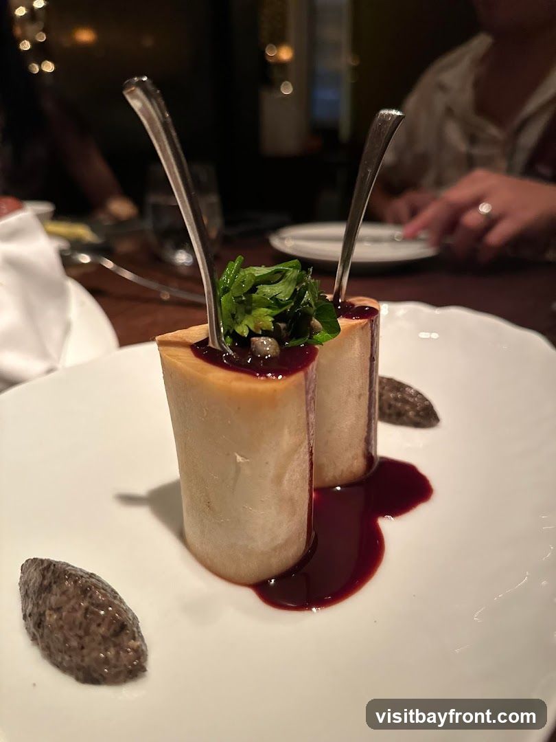 A close-up shot of two pieces of roasted bone marrow, each topped with fresh herbs and capers, served on a white plate with a dark red sauce and a side of what appears to be a mushroom puree. Forks are inserted into the bone marrow. The background is blurred, showing a dimly lit dining setting with other patrons.