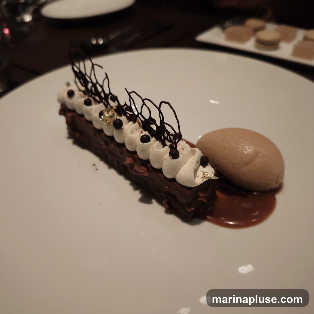 A chocolate dessert bar topped with whipped cream, chocolate shavings, and gold leaf, served with a scoop of ice cream and caramel sauce on a white plate.