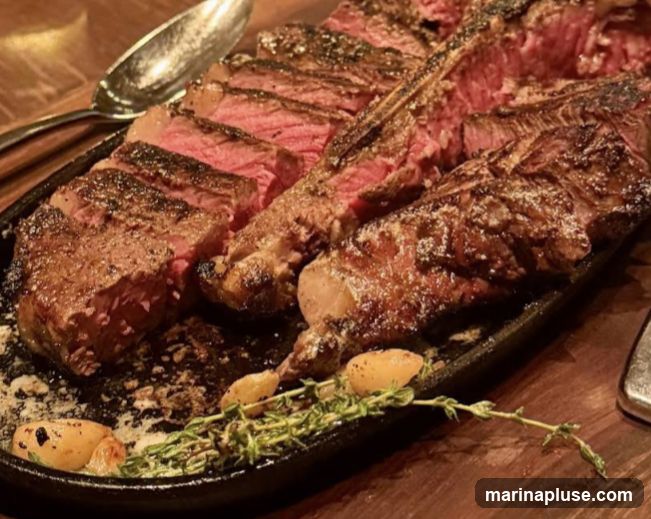 A close-up shot of a sliced porterhouse steak served on a cast-iron platter. The steak is cooked to a medium-rare, with a pink interior and a well-seared crust. It is garnished with roasted garlic cloves and fresh thyme sprigs, and appears to be seasoned with salt and pepper. A large serving spoon is visible to the left of the steak. The platter rests on a wooden surface.
