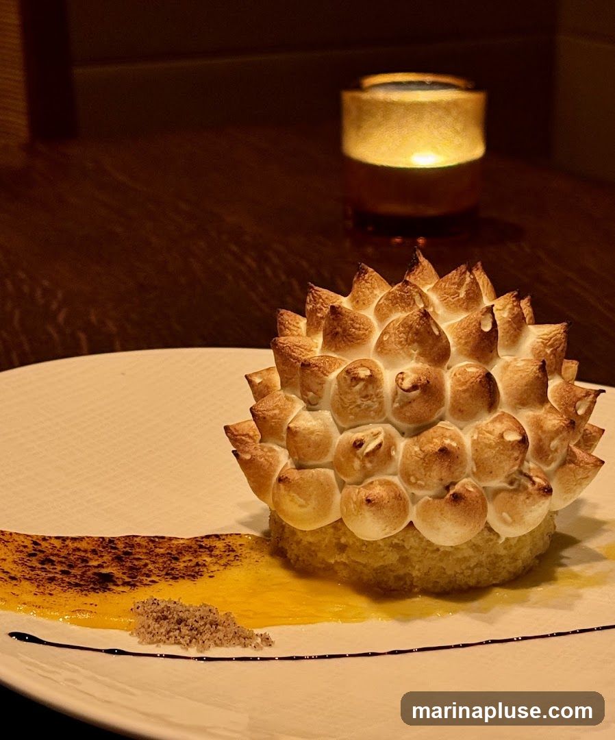 A close-up shot of a dessert on a white plate. The dessert is a small cake topped with peaks of torched meringue, resembling a spiky fruit. It is served with a swirl of yellow sauce and a sprinkle of nuts. A lit candle in a glass holder is visible in the blurred background.