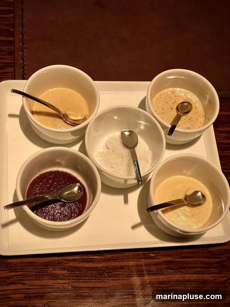 A white tray holds five small white bowls, each with a spoon. Three bowls contain a light-colored, creamy dessert. One bowl has a dark red, jam-like substance. Another bowl contains white granular material, possibly sugar or salt. The tray is placed on a dark wooden surface.