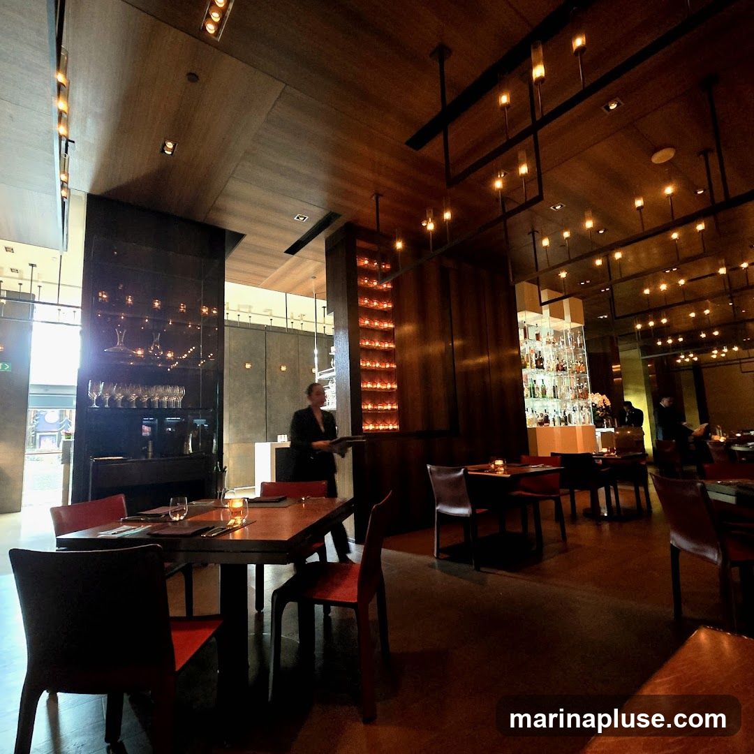 A dimly lit, upscale restaurant interior with wooden paneling and modern lighting fixtures. Tables are set with red chairs and place settings. A server in a black suit is visible carrying a tray. A well-stocked bar is in the background.