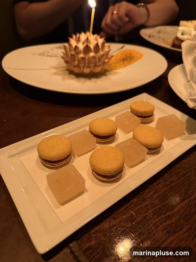 A white rectangular platter holds several yellow macarons and clear, cubed jellies. In the background, a round dessert with meringue peaks is topped with a lit birthday candle. The dessert sits on a white plate with some writing on it and a smear of orange powder. Another plate with dessert is visible in the far background. The setting appears to be a restaurant with dark wood tables.