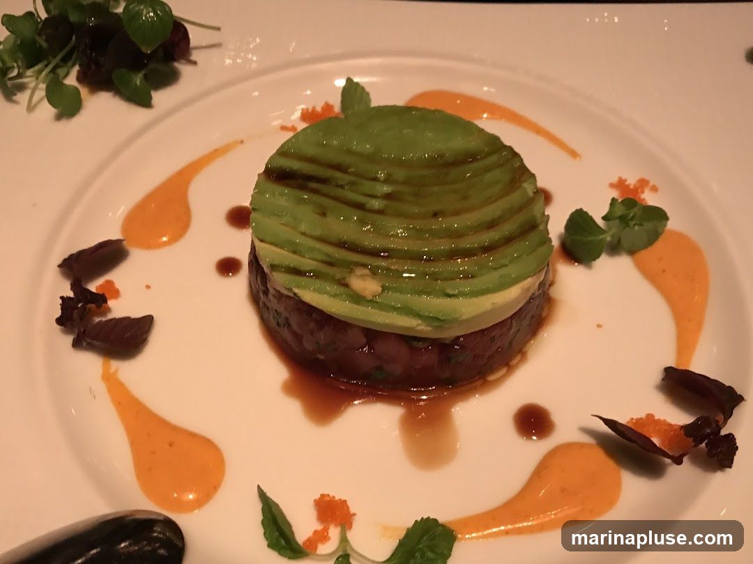 A close-up shot of a tuna tartare dish, presented on a white plate. The tartare is topped with thinly sliced avocado, drizzled with a dark sauce. The plate is garnished with dollops of orange sauce, microgreens, and small pieces of dark, crispy garnish. A sprig of greens and some dark leaves are visible in the upper left corner.