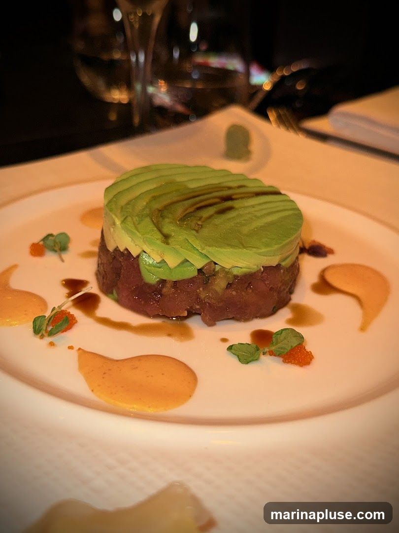 A close-up shot of a tuna tartare dish, topped with thinly sliced avocado. The dish is served on a white plate with dollops of orange sauce and garnished with microgreens and small orange roe. A glass of white wine and a fork are visible in the blurred background.