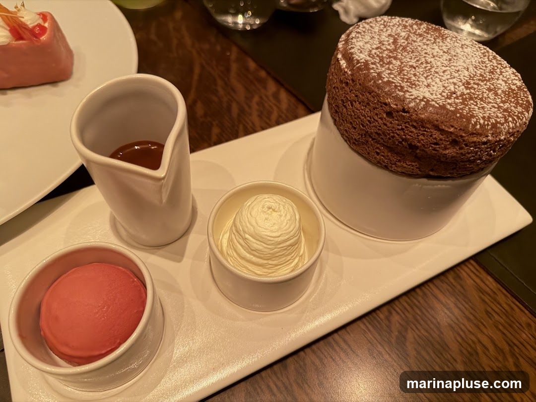 A white platter holds a chocolate souffle dusted with powdered sugar, a scoop of whipped cream, a scoop of pink sorbet or ice cream, and a small pitcher of caramel sauce. A slice of pink cake with white frosting is visible on a white plate in the upper left corner.
