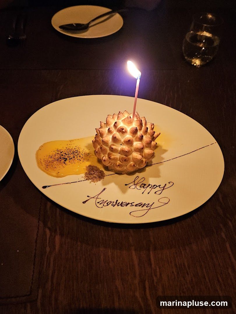 A dessert with a lit candle sits on a white plate. The dessert is topped with meringue and appears to be shaped like a pinecone. It is served with a yellow sauce and sprinkled with dark powder. The plate is decorated with the words "Happy Anniversary" written in dark icing. The setting appears to be a restaurant with dark wood tables.