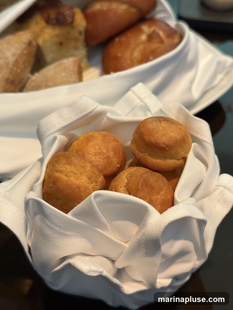 A close-up shot of a white cloth-lined basket filled with small, golden-brown rolls. In the background, another basket holds larger bread pieces and rolls.