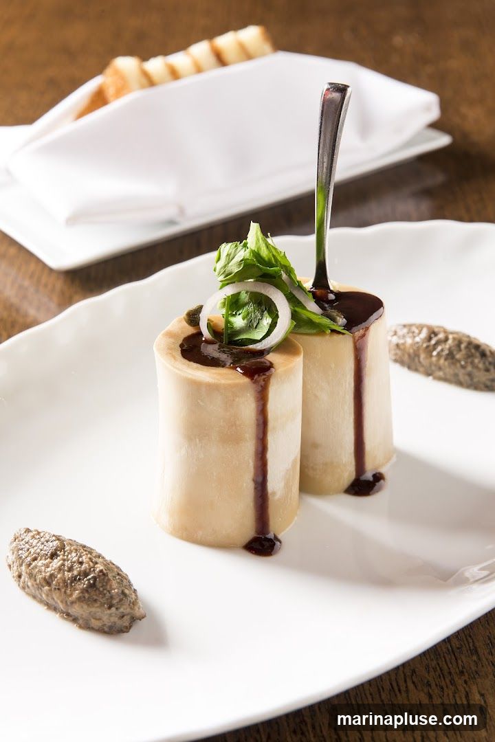 A close-up shot of a gourmet appetizer featuring two pieces of bone marrow, drizzled with a dark sauce and garnished with fresh herbs and a slice of onion. Two small quenelles of a pâté-like spread are placed on either side of the bone marrow. In the background, toasted bread slices are neatly arranged on a white napkin.