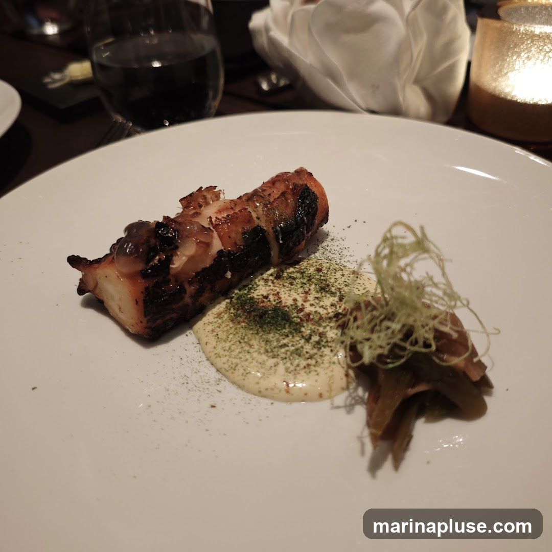 A close-up shot of a piece of grilled octopus served on a white plate. The octopus is charred in places and appears tender. It is accompanied by a dollop of creamy sauce, sprinkled with green herbs and spices. To the right of the sauce is a small portion of pickled vegetables or garnish. In the background, a glass of dark liquid, a folded white napkin, and a lit candle in a glass holder are visible, suggesting a restaurant setting.