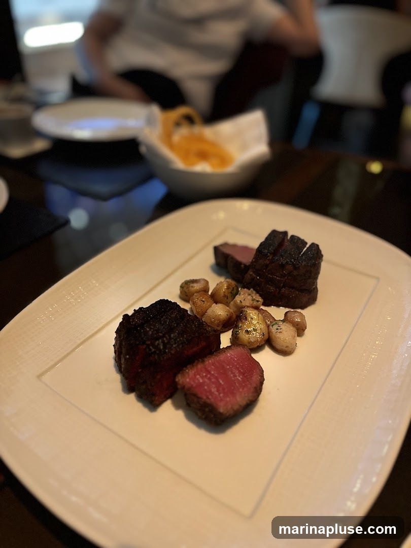 A plate of steak and potatoes is presented on a white textured plate. The steak is cut into several pieces, some whole and some sliced to reveal a medium-rare interior. Small, roasted potatoes are scattered around the steak. In the background, out of focus, are diners and a bowl of what appears to be onion rings.