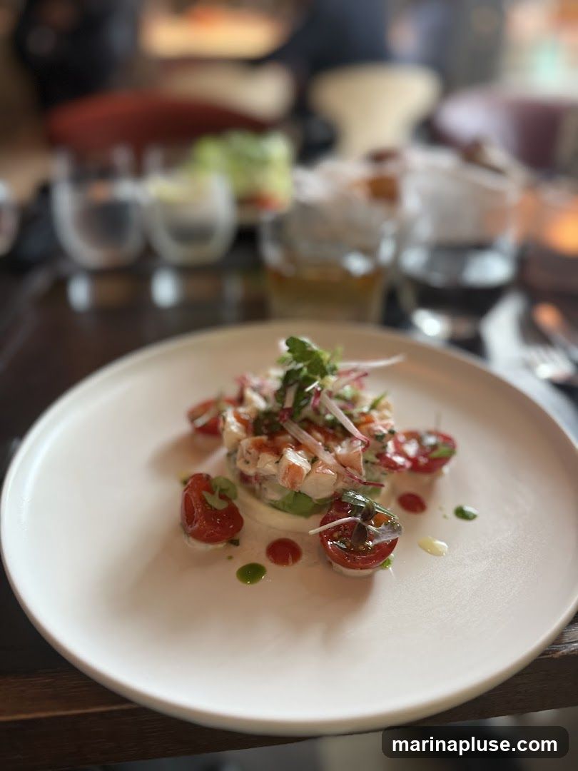 A close-up shot of a delicate seafood salad served on a white plate. The salad is topped with fresh herbs and thinly sliced red onions, and is garnished with halved cherry tomatoes and a drizzle of green sauce. In the blurred background, glasses of drinks and other diners are visible, suggesting a restaurant setting.