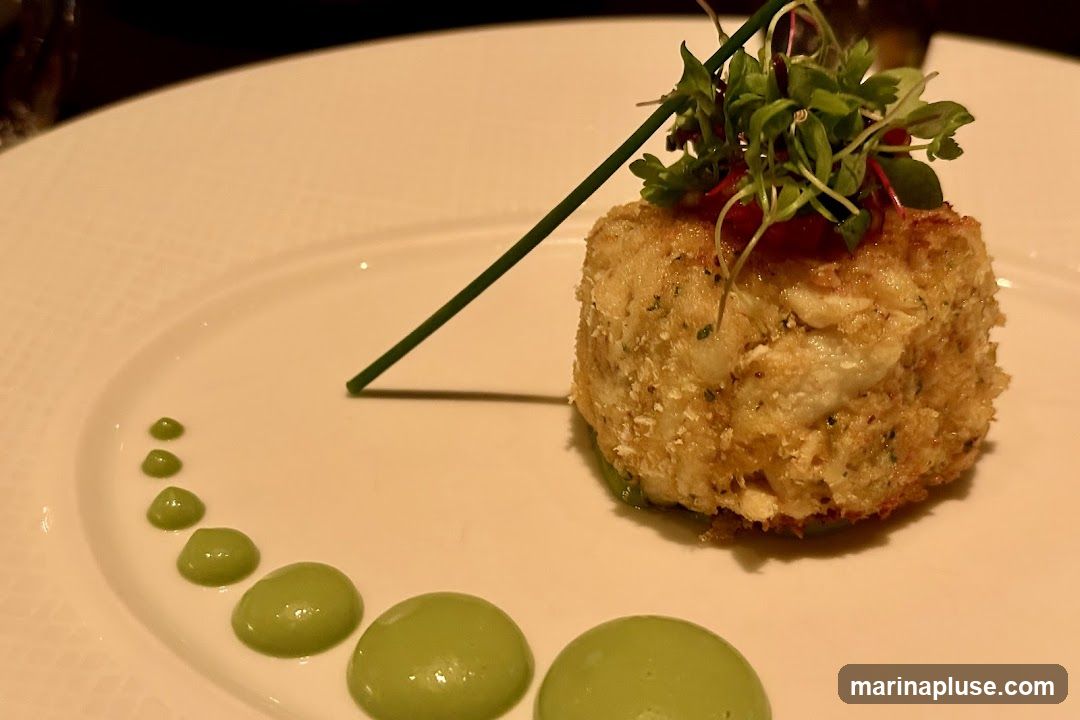 A close-up shot of a golden-brown crab cake on a white plate. The crab cake is topped with fresh microgreens and a thin red garnish. A chive stalk is placed diagonally across the crab cake. A series of small, vibrant green dots of sauce are artfully arranged in a curve on the left side of the plate.