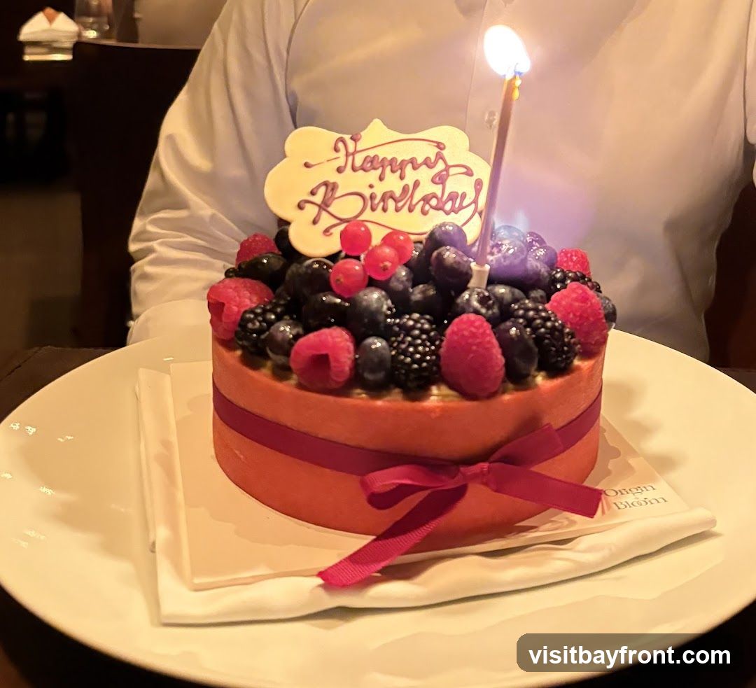 A round, orange-frosted cake topped with a variety of fresh berries including blueberries, raspberries, blackberries, and red currants. A lit birthday candle stands in the center, and a white plaque with "Happy Birthday" written in purple icing is placed on top. The cake is tied with a dark pink ribbon and sits on a white plate with a white napkin underneath. The text "Origin Blom" is visible on the napkin. A person in a white shirt is partially visible in the background.