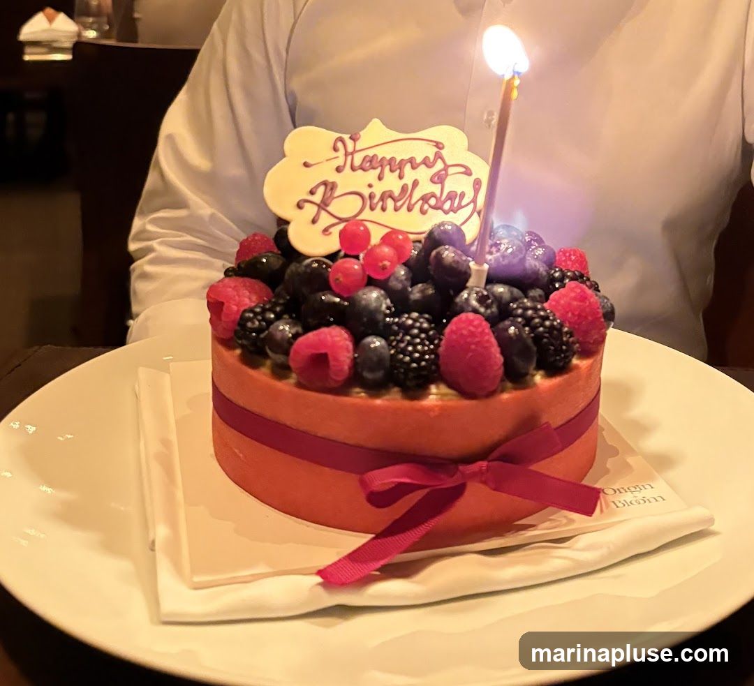 A round, orange-frosted cake topped with a variety of fresh berries including blueberries, raspberries, blackberries, and red currants. A lit birthday candle stands in the center, and a white plaque with "Happy Birthday" written in purple icing is placed on top. The cake is tied with a dark pink ribbon and sits on a white plate with a white napkin underneath. The text "Origin Blom" is visible on the napkin. A person in a white shirt is partially visible in the background.