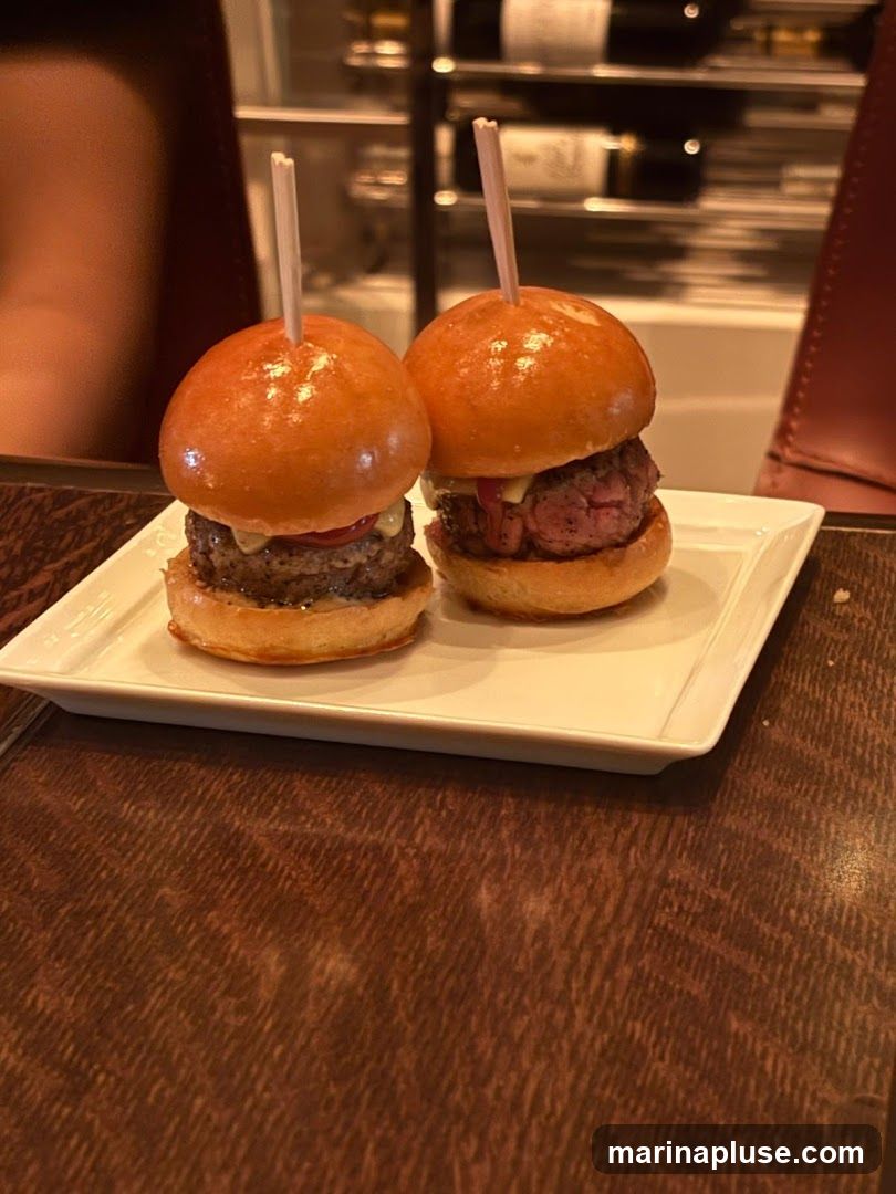 Two sliders are presented on a white rectangular plate. Each slider has a glossy bun, a thick beef patty, and a toothpick inserted through the top. The sliders appear to be garnished with some sauce or relish. The plate sits on a dark wooden table. In the background, out of focus, are shelves with bottles and chairs.