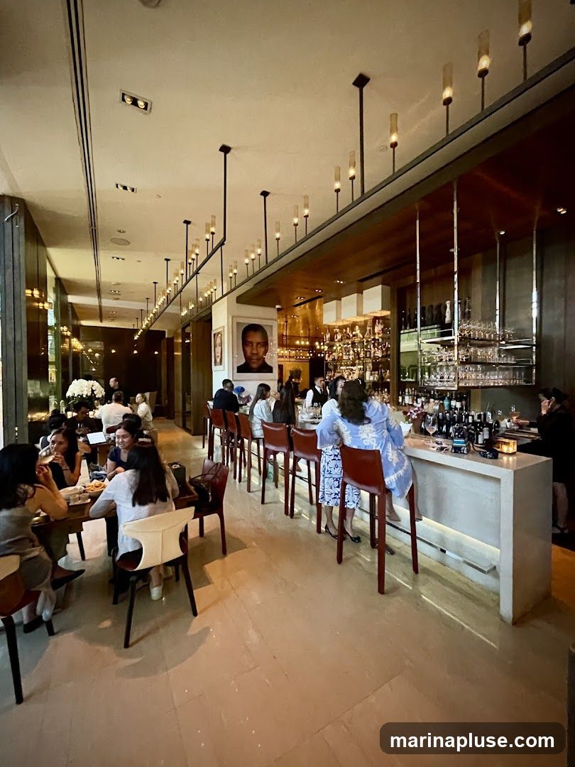 The image shows the interior of a restaurant with a long bar and dining tables. Several patrons are seated at the tables and at the bar. The bar area is well-lit with bottles of alcohol and glassware displayed on shelves behind the bar. A portrait of a man hangs on the wall above the bar.