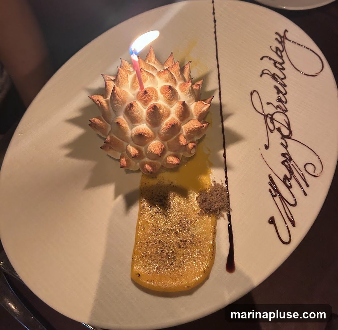 A dessert on a white plate with a lit birthday candle on top of a meringue-like topping. The dessert also has a yellow sauce and a sprinkle of brown crumbs. The words "Happy Birthday" are written on the plate in chocolate sauce.