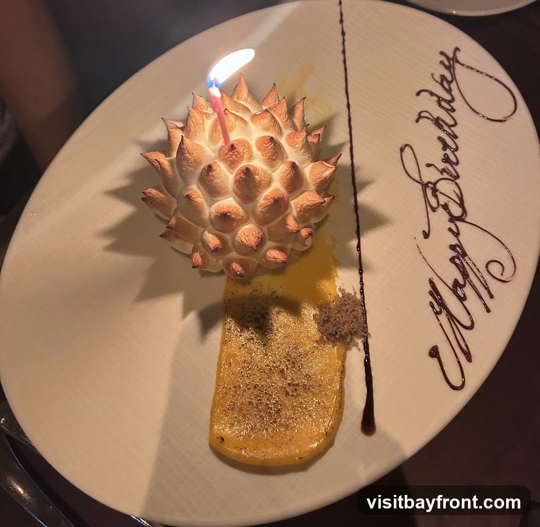 A dessert on a white plate with a lit birthday candle on top of a meringue-like topping. The dessert also has a yellow sauce and a sprinkle of brown crumbs. The words "Happy Birthday" are written on the plate in chocolate sauce.