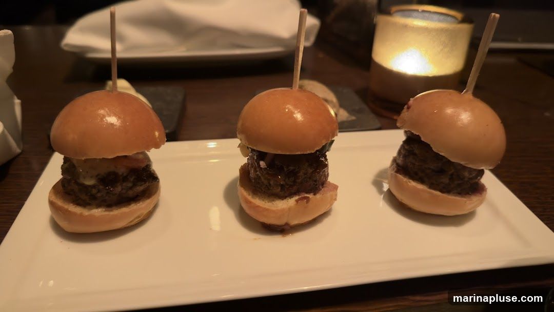 Three mini burgers, each with a toothpick inserted, are arranged on a rectangular white plate. The burgers have a meat patty and appear to have various toppings. In the background, a lit candle in a holder and some white napkins are visible. The setting suggests a restaurant or dining environment.