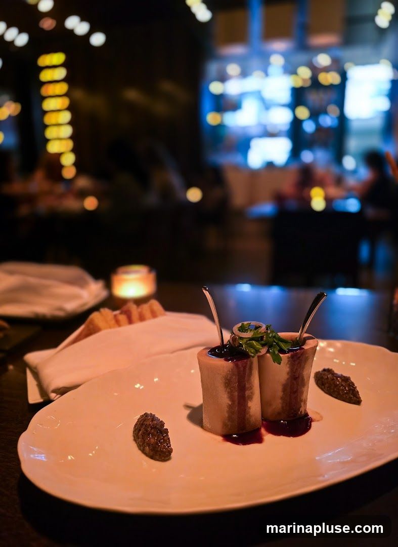 A close-up shot of a gourmet appetizer featuring two pieces of bone marrow, served upright on a white oval plate. The marrow is topped with a dark sauce and garnished with fresh parsley and a ring of thinly sliced onion. Two dollops of a dark, textured relish or spread are placed on either side of the marrow. In the background, a white napkin-wrapped bread basket and a lit candle are visible, along with the blurred ambiance of a dimly lit restaurant interior with patrons and decorative lighting.